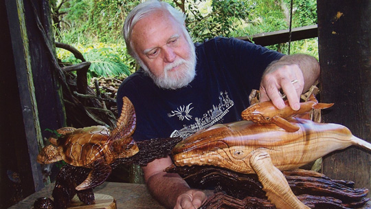 an elderly man with white hair looks at wooden sculptures of a humpback whale and sea turtles while sitting in his backyard studio set against a forest backdrop