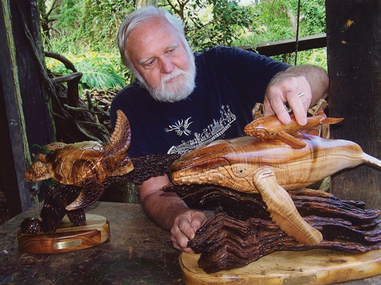 an elderly man with white hair looks at wooden sculptures of a humpback whale and sea turtles while sitting in his backyard studio set against a forest backdrop