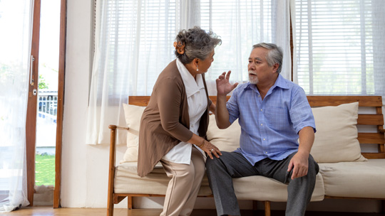 an elderly couple have a worried conversation on a couch in a bright and airy living room