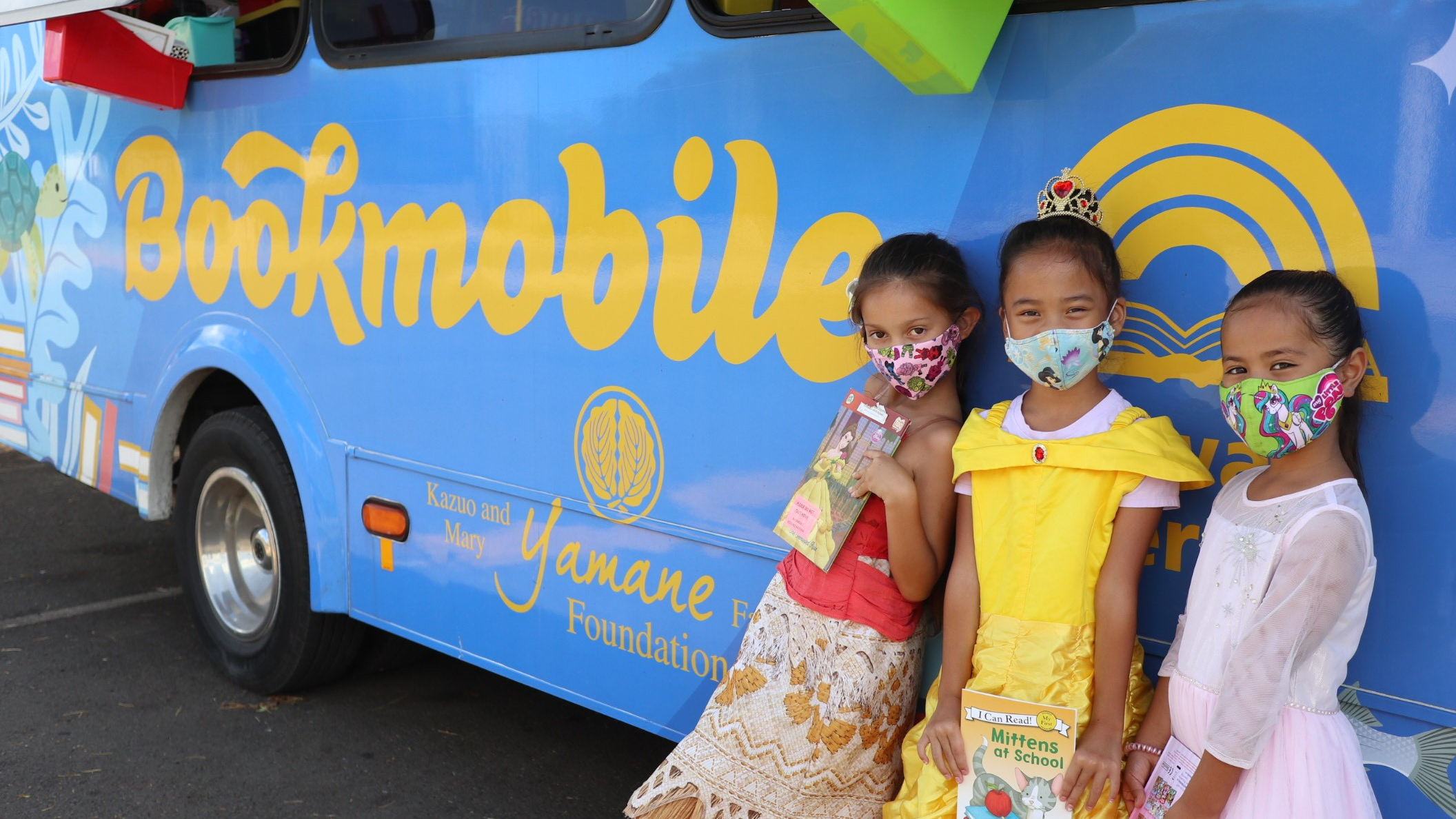 three girls dressed in Disney princess costumes stand against the blue Bookmobile bus and hold books in their hands