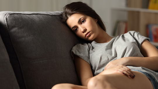 a young woman sits on a couch with her arms wrapped around her body and looking off into the distance with a worried expression on her face