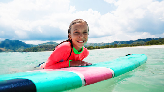 young girl in a neon rash guard floating next to a surfboard in the ocean with the Koolau mountains in the background