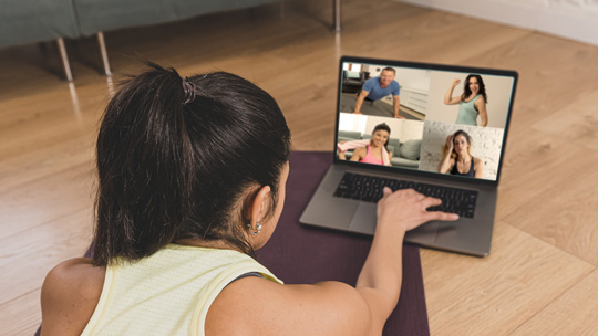 a young woman in workout clothes logs onto a laptop computer set up on a yoga mat on the living room floor. her computer screen displays a man and three women ready to workout