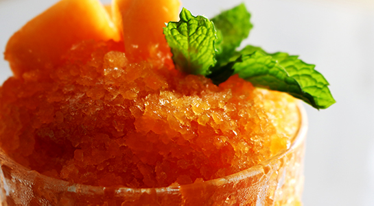 a close-up side view of a deep-orange cantaloupe papaya granita served in an etched glass topped with green mint sprigs and light orange cantaloupe chunks