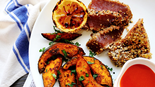 a close-up view of seared ahi crusted with panko and sesame seeds next to roasted kabocha squash wedges, a small dish of red hot sauce, and a broiled lemon wedge