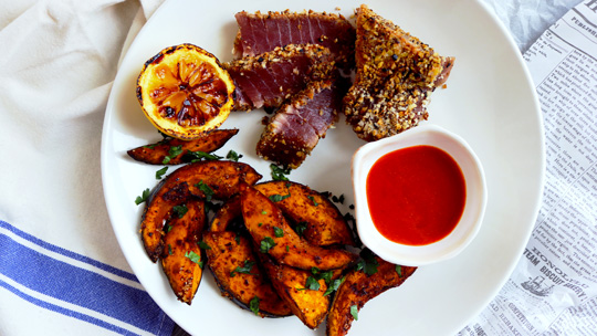 an overhead view of seared ahi crusted with panko and sesame seeds next to roasted kabocha squash wedges, a small dish of red hot sauce, and a broiled lemon on a plate atop a blue and white napkin and crumpled newspaper wedge