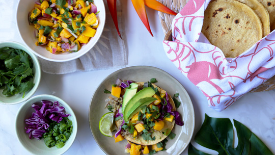 a plate of freshly made kalua pork carnitas sits on a white marble counter surrounded by a big bowl of mango salsa, a platter of charred corn tortillas and tropical flowers
