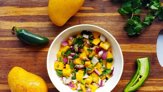 a bowl of mango salsa sits on a wooden cutting board surrounded by mangos cilantro and jalapenos
