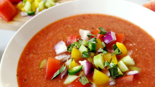 close-up of a bowl of watermelon and cucumber gazpacho topped with cubed yellow bell peppers, cucumbers, red onion, and green peppers with a cutting board with more cubed vegetables and watermelon in the background
